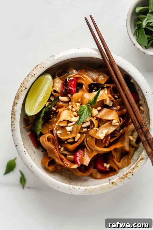 A close-up shot of Thai Drunken Noodles in a white ceramic bowl, showcasing wide rice noodles, fresh Thai basil, and chili peppers.
