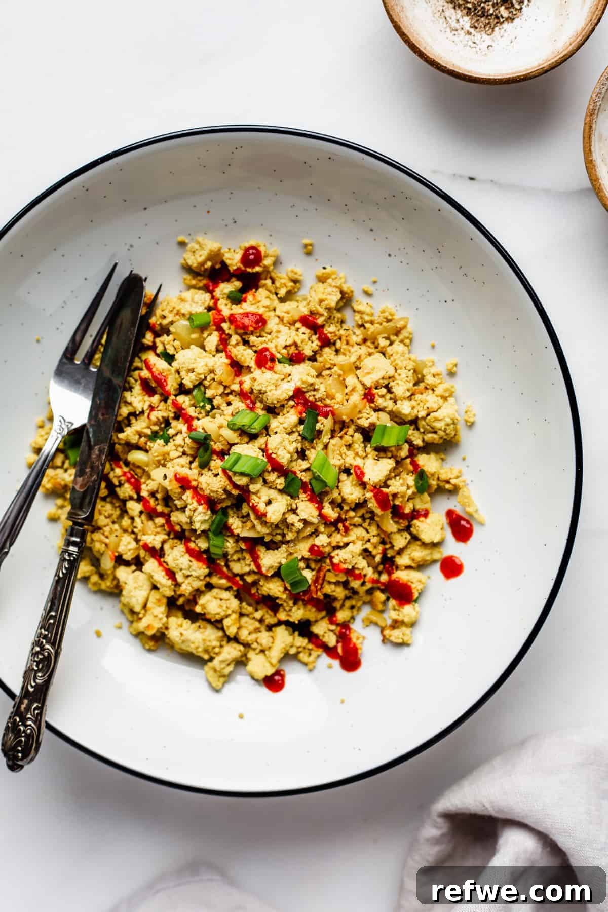 Next-Level Tofu Scramble 2 A vibrant bowl of fluffy scrambled tofu, generously topped with freshly chopped green onions and a drizzle of spicy sriracha, ready to be enjoyed as a healthy vegan breakfast.