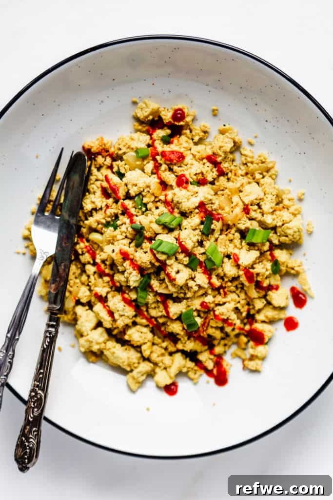 Next-Level Tofu Scramble 6 A vibrant and beautifully plated scrambled tofu dish, garnished with fresh green onions and a swirl of sriracha, in an elegant white bowl with a subtle black rim.