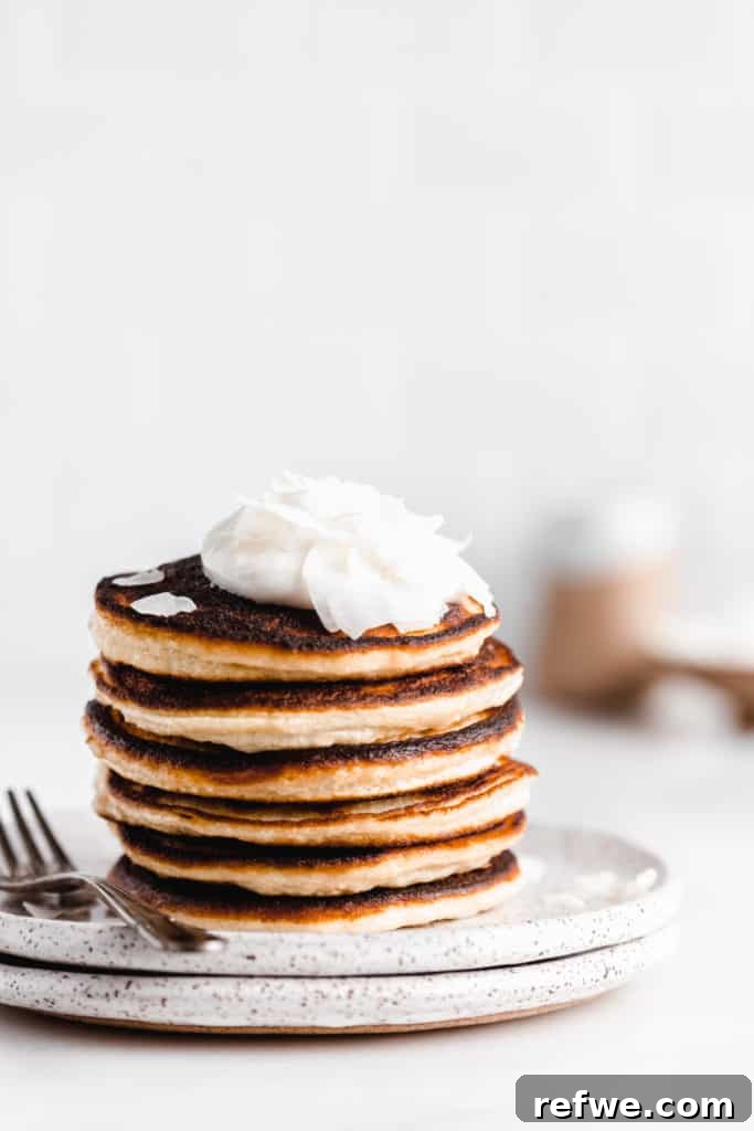 a stack of coconut flour pancakes topped with yogurt and coconut flakes with maple syrup being drizzled on top