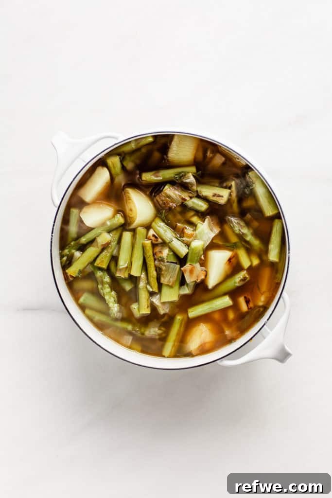 Leeks, asparagus, potato, and vegetable broth simmering in a white pot with fresh herbs.