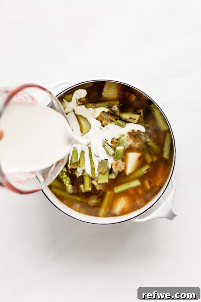 A measuring cup pouring smooth cashew cream into a pot of simmering vegetable and asparagus soup.