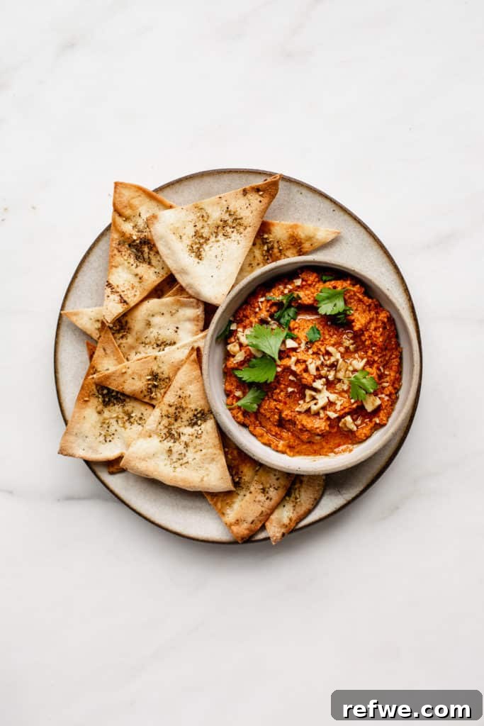 Roasted Red Pepper Walnut Muhammara 2 A ceramic plate with pita chips on it and a bowl of roasted red pepper dip, garnished with parsley and crushed walnuts.
