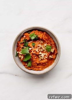 A ceramic white bowl with roasted red pepper dip topped with crushed walnuts and parsley