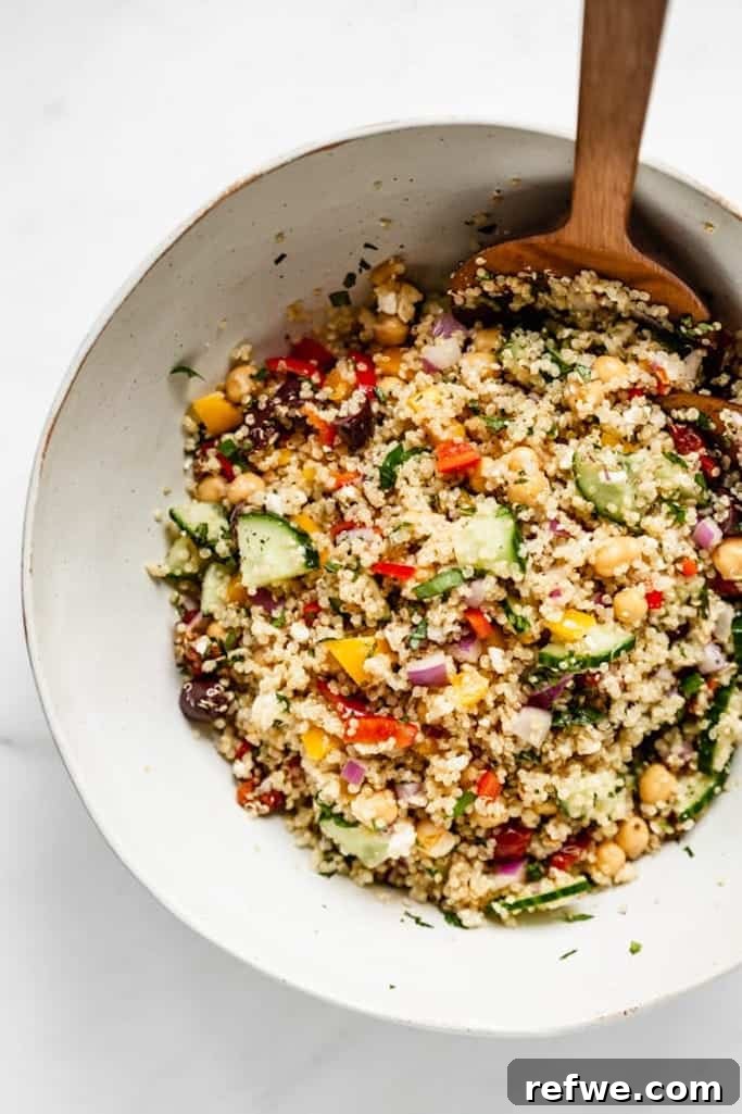 white ceramic bowl filled with summer salads made of quinoa, cucumbers, red and yellow peppers, with a large wooden spoon