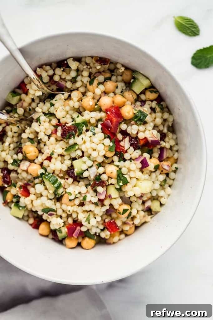 white serving bowl filled with israeli couscous mixed with fresh vegetables and herbs and a silver spoon