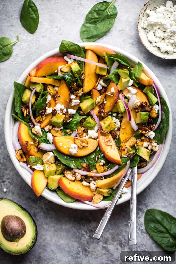 summer peach spinach salad with sliced peaches, red onions and toasted almonds in a large white bowl with chopped avocado and silver utensils, against a grey background