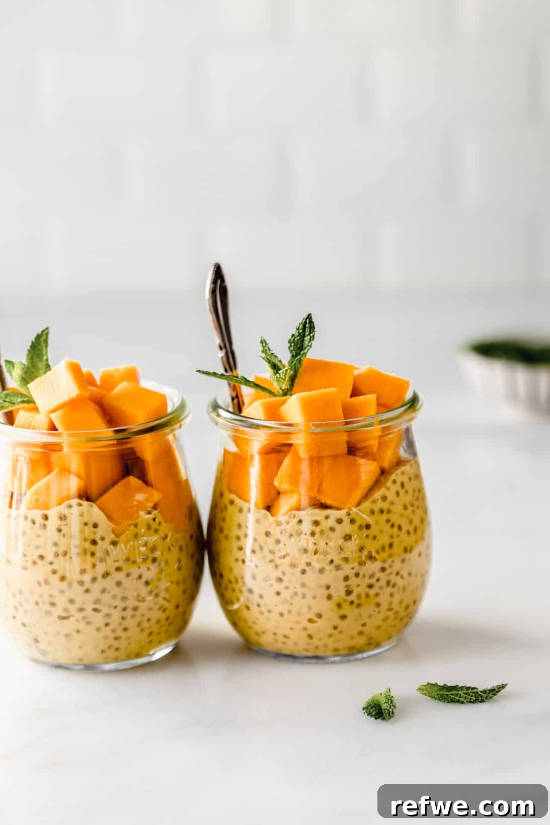Two jars of mango chia pudding, garnished with fresh mango cubes and mint, placed on a rustic wooden board.