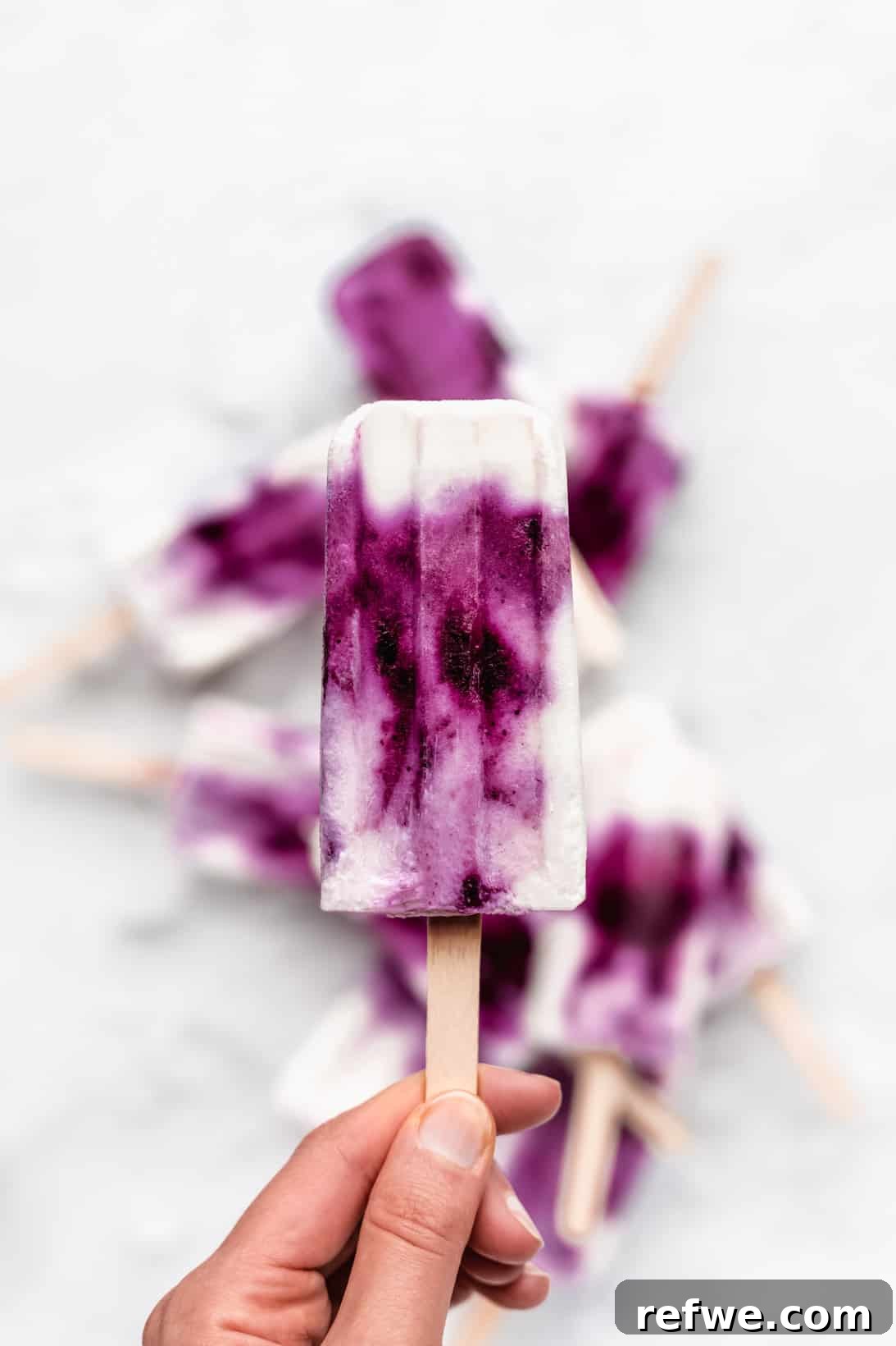 A hand gracefully holding a single blueberry Greek yogurt popsicle against a soft, bright background