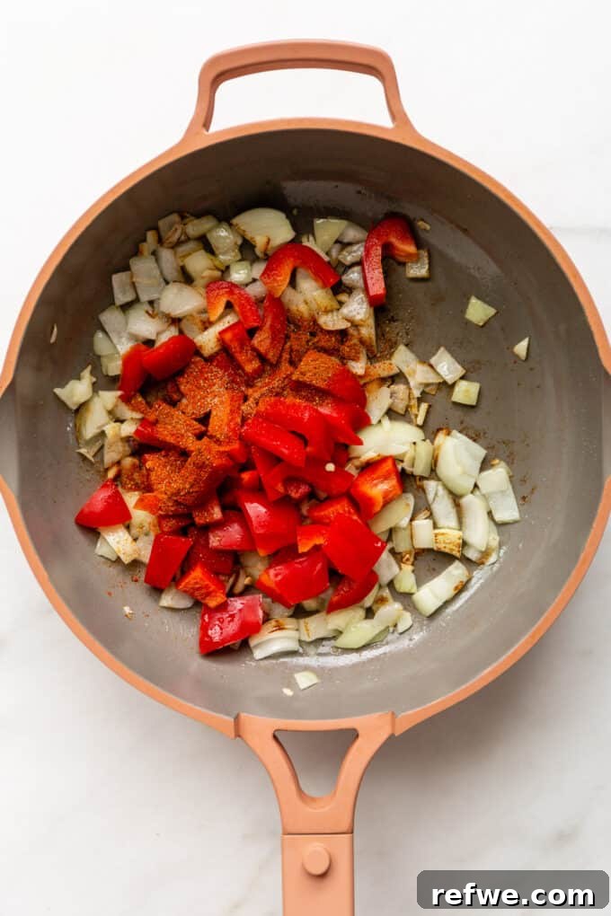 A pink skillet filled with chopped onions, bell peppers, and various spices, ready to be sautéed.