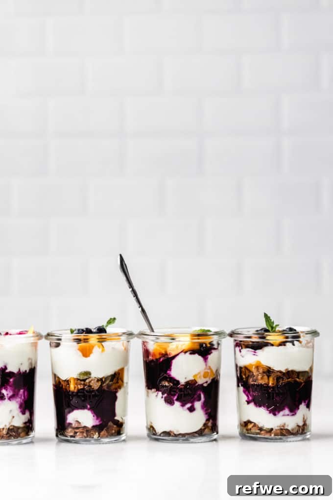 Four artistic jars of yogurt and granola parfaits, beautifully adorned with blueberries, mango, and fresh mint leaves, showcasing their vibrant layers.