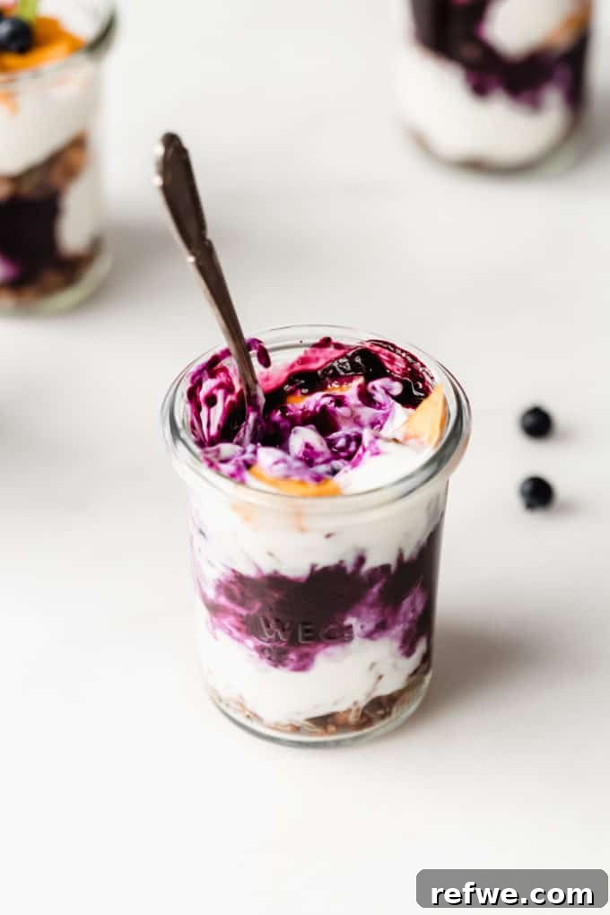 A half-assembled jar of yogurt parfait, showing distinct layers of creamy yogurt, bright blueberry compote, and fresh mango cubes, enticingly ready for more.