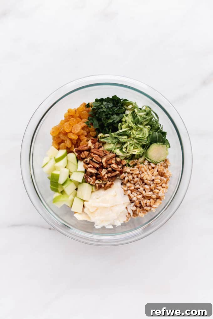 Farro, Brussels sprouts, parsley, golden raisins, apples, Parmesan cheese, and pecans arranged in a clear mixing bowl, ready to be combined.