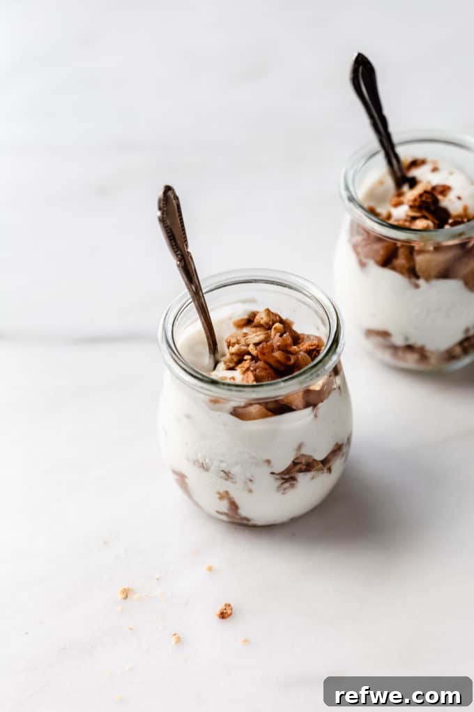 Cinnamon Apple Parfait Stacks 2 Two glass jars filled with layered apple pie parfaits, showing stewed apples, crumble, and yogurt, with spoons resting inside.