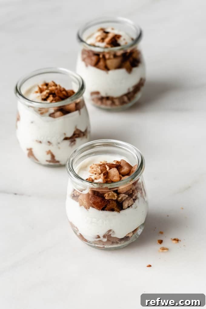 Cinnamon Apple Parfait Stacks 6 Three beautifully layered apple pie parfaits in glass jars, displaying alternating layers of creamy yogurt, golden granola, and luscious cooked apples.