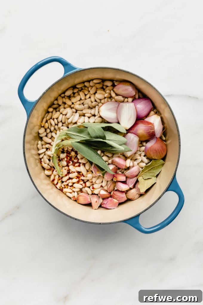 Oven Braised Beans 3 A blue pot showcasing all the raw ingredients for brothy beans: white cannellini beans, whole garlic bulbs, shallots, fresh sage, and bay leaves, submerged in vegetable stock.