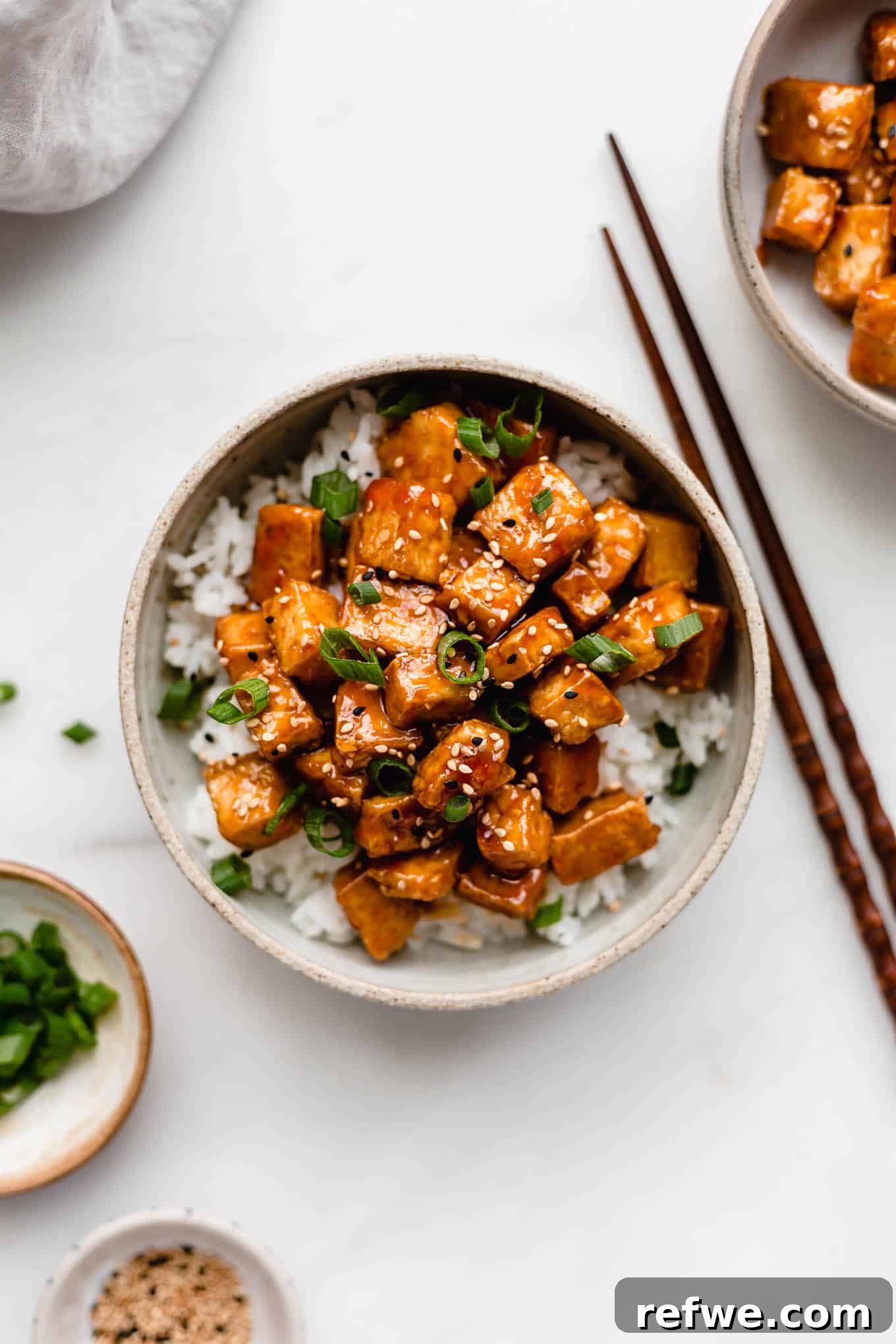 Caramelized Tofu 2 Sweet and sour tofu in a white ceramic bowl with two small bowls on the side filled with green onions and sesame seeds
