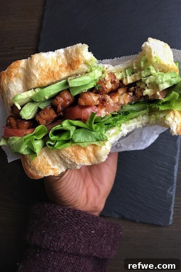 Unlock Tempeh's Potential 23 Recipes and a Full Cooking Guide 22 A hand presenting a perfectly layered Vegan BLT sandwich, featuring sweet and savory tempeh bacon, crisp lettuce, fresh tomato, and creamy avocado.