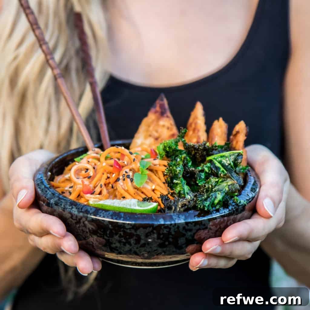 Unlock Tempeh's Potential 23 Recipes and a Full Cooking Guide 5 A hand holding a black bowl filled with creamy sweet potato noodles, crispy garlic tempeh, and vibrant kale.