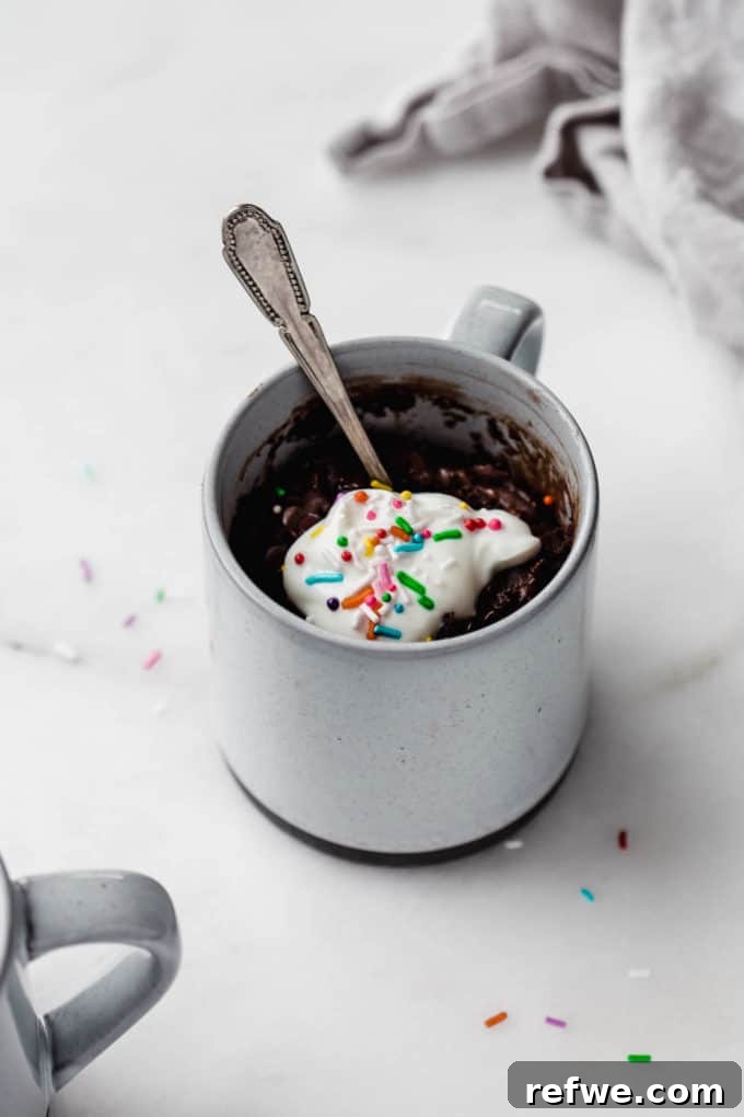 A perfectly baked vegan chocolate mug cake in a stylish grey mug, adorned with light whipped cream and cheerful rainbow sprinkles.