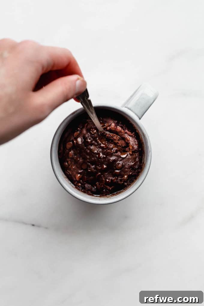 A freshly microwaved vegan chocolate mug cake in a grey mug, with a spoon inserted, ready to be enjoyed warm.