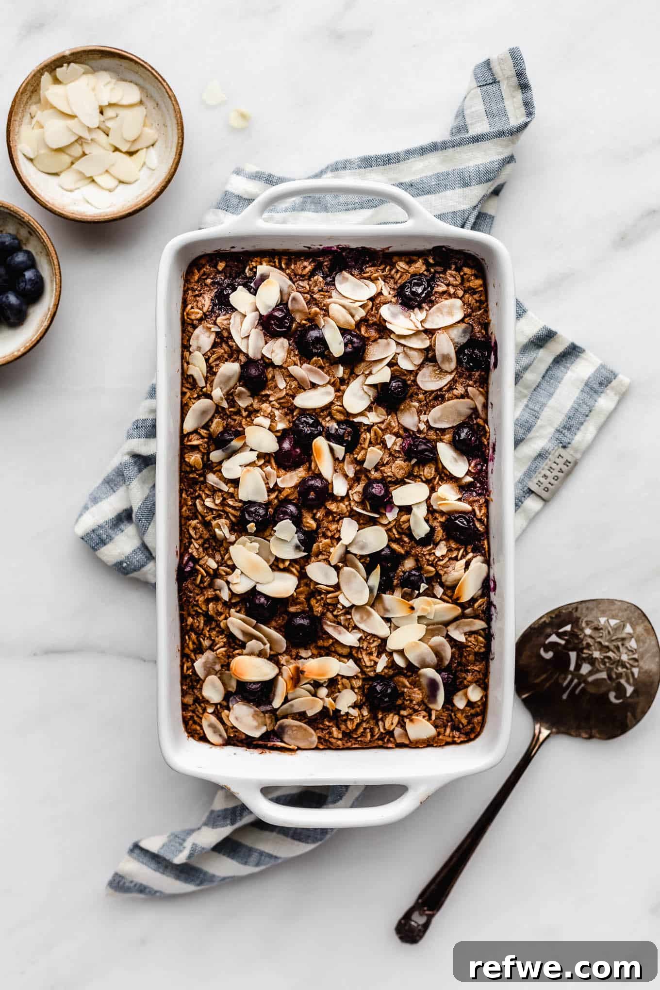 A baking dish with blueberry baked oatmeal in it on a blue napkin, garnished with fresh blueberries and a light drizzle