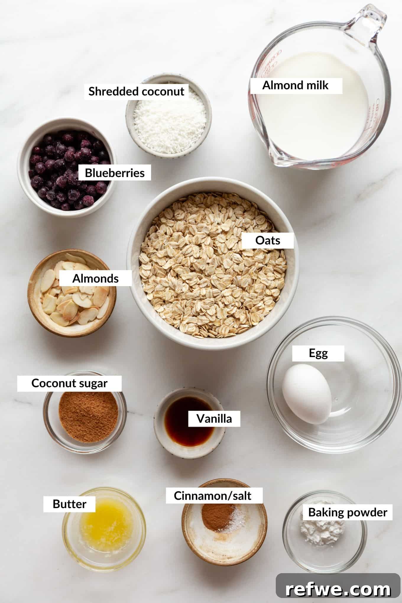 Ingredients for blueberry baked oatmeal laid out on a white surface, including oats, blueberries, milk, eggs, and spices.