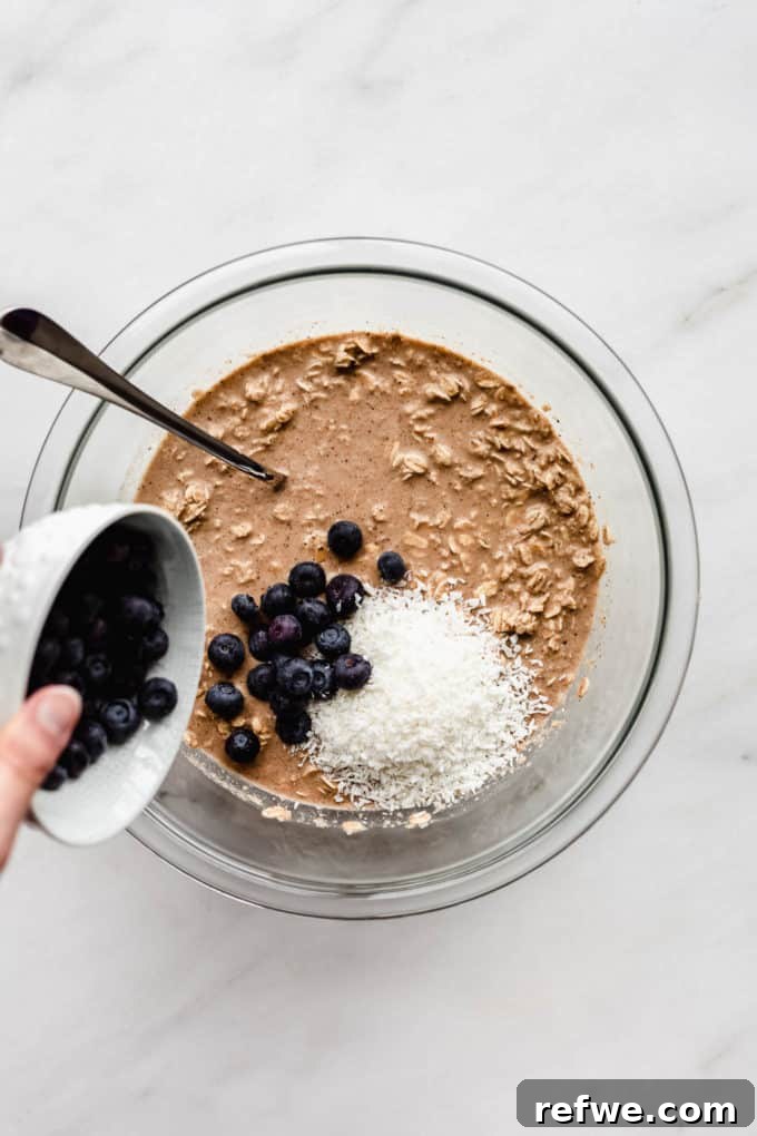 A cup of fresh blueberries being poured into a mixing bowl of baked oatmeal batter.