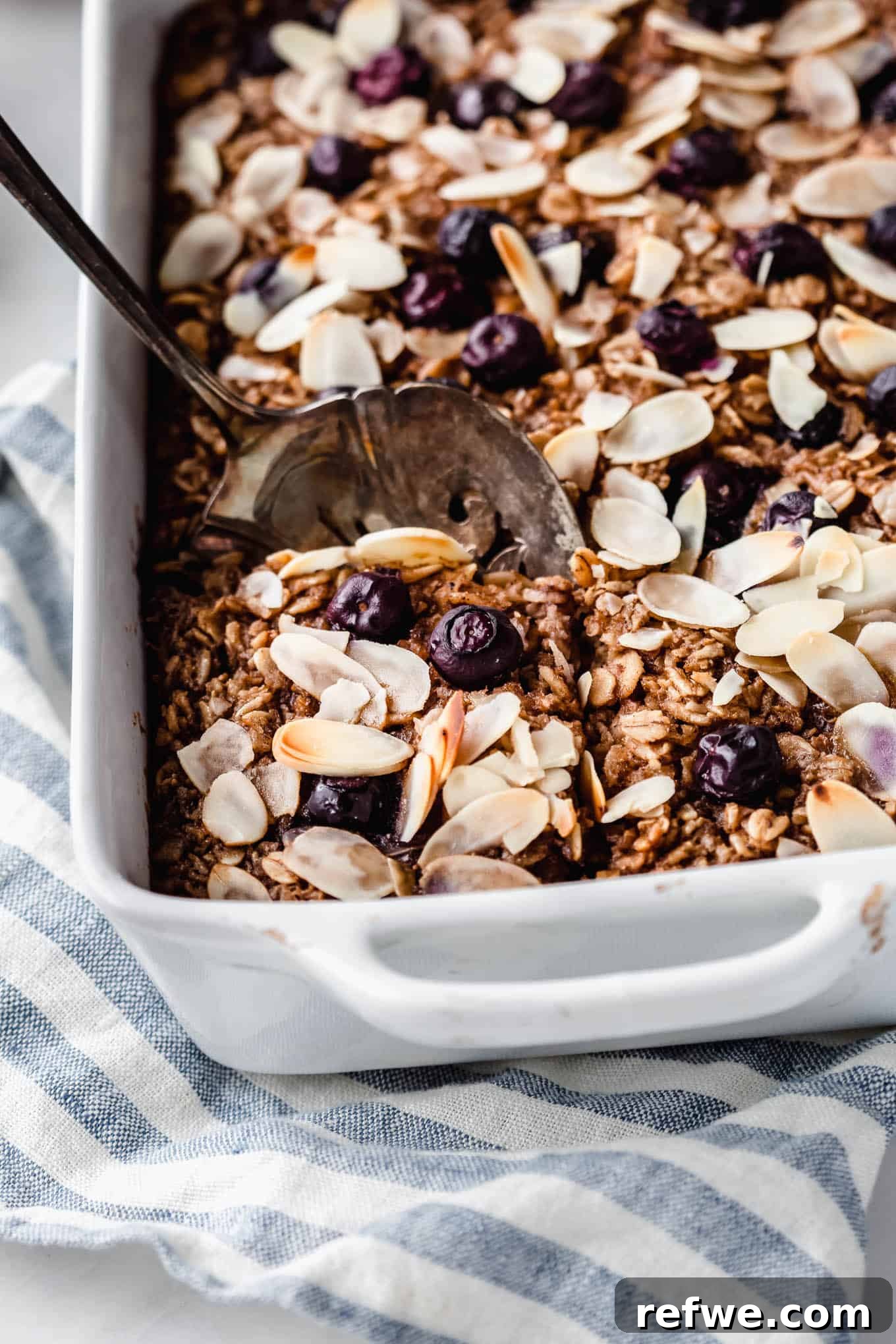 A spoon scooping a portion of blueberry baked oatmeal from a white baking dish, showing the moist, fruity interior.