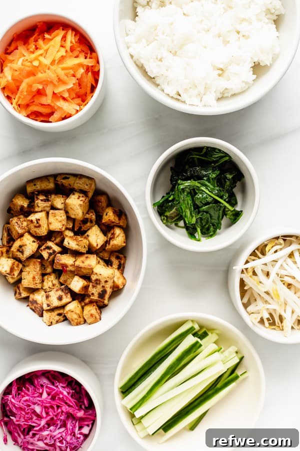 A flat lay photograph showcasing the individual ingredients prepared for a vegan bibimbap bowl: cooked rice, crispy tofu, pickled cabbage, grated carrots, sliced cucumber, sautéed spinach, and a jar of sauce.