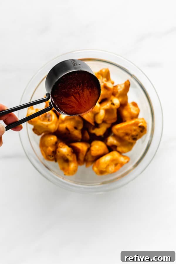 A measuring cup pouring buffalo sauce into a bowl of crispy cauliflower wings