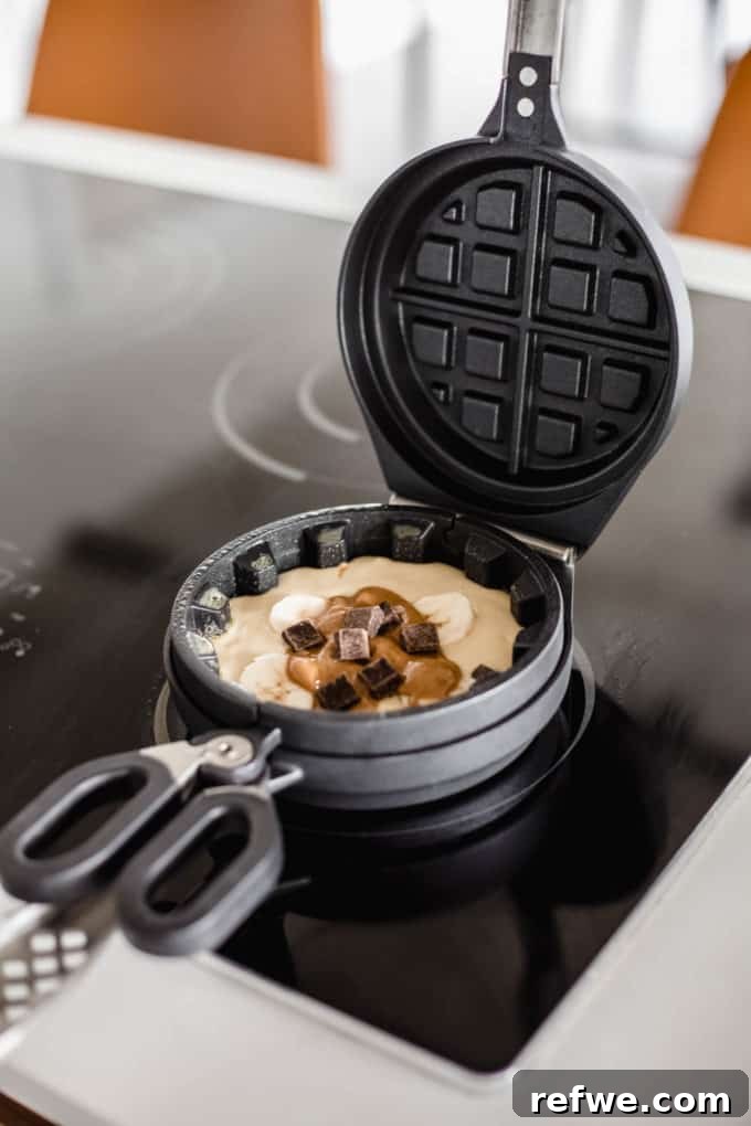 Snickers Banana Bliss Waffles 5 The Wonderffle waffle iron with a layer of batter, topped with banana slices, dollops of peanut butter, and a generous sprinkle of dark chocolate chips.