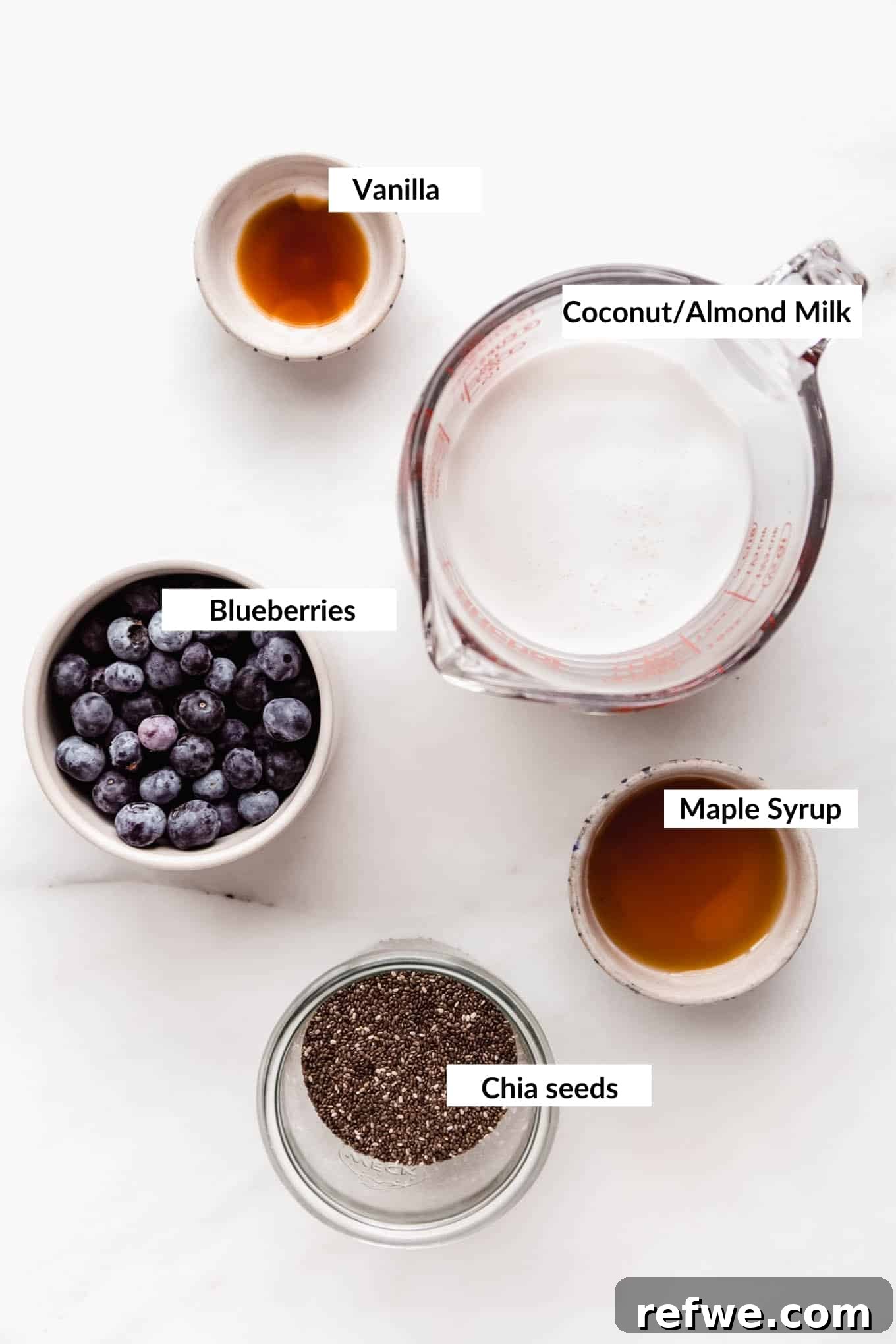 A flat lay photograph showcasing the key ingredients laid out neatly on a surface: fresh blueberries, chia seeds, a bottle of plant-based milk, and a small container of maple syrup, all ready for making blueberry chia pudding.