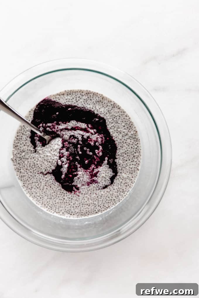 A close-up shot of a clear mixing bowl containing a creamy chia pudding mixture, with a distinct layer of freshly cooked blueberry compote being added on top, ready for mixing.
