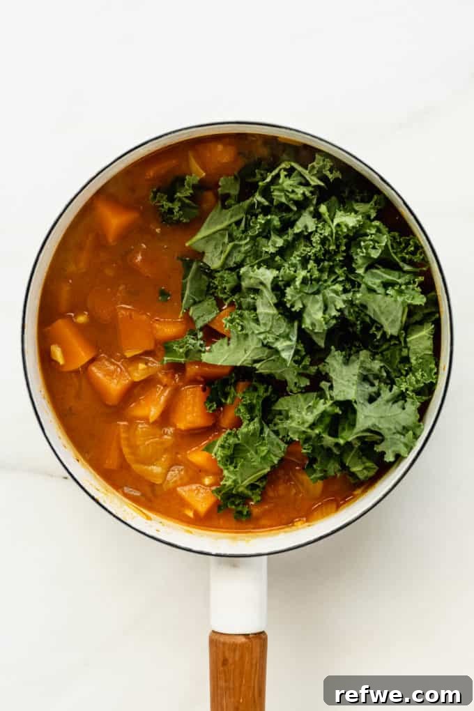 Velvet Squash Curry 8 A white pot with butternut squash curry and fresh kale leaves being added