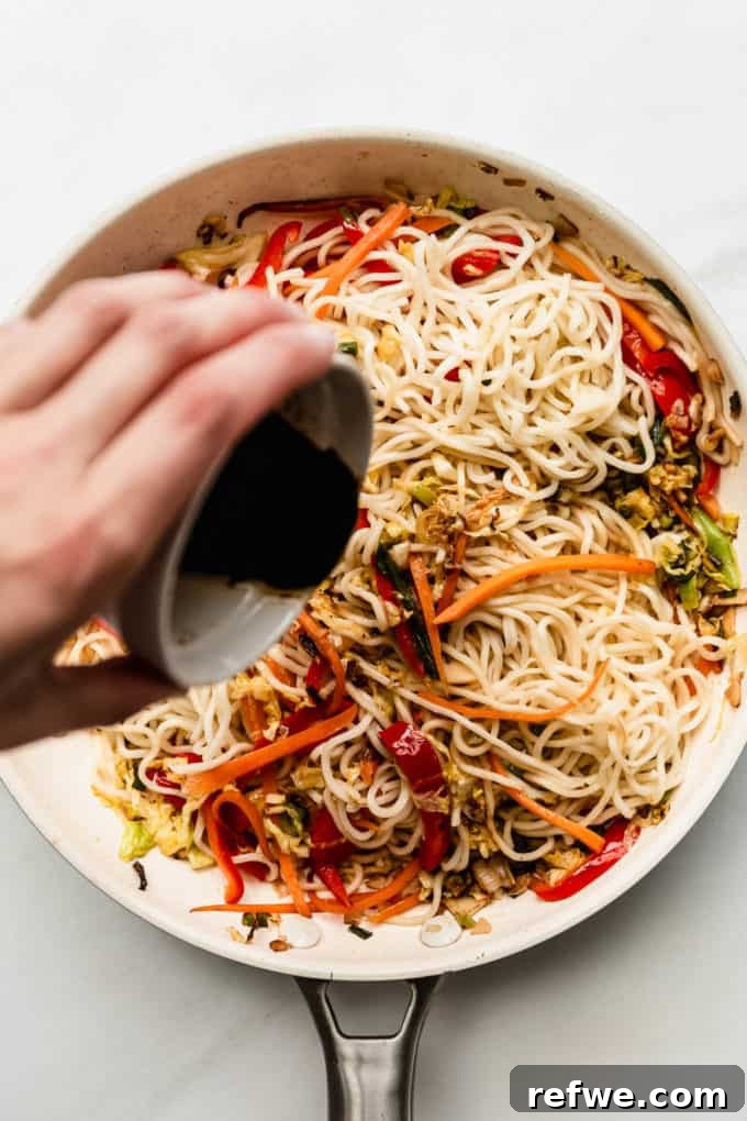 Plant-Based Vegetable Noodle Stir-Fry 7 A hand is captured pouring the rich, dark stir-fry sauce over the noodles and vegetables in the pan.