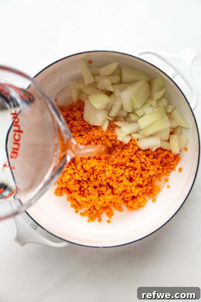 A clear measuring cup carefully pouring water into the pot with red lentils and onions.