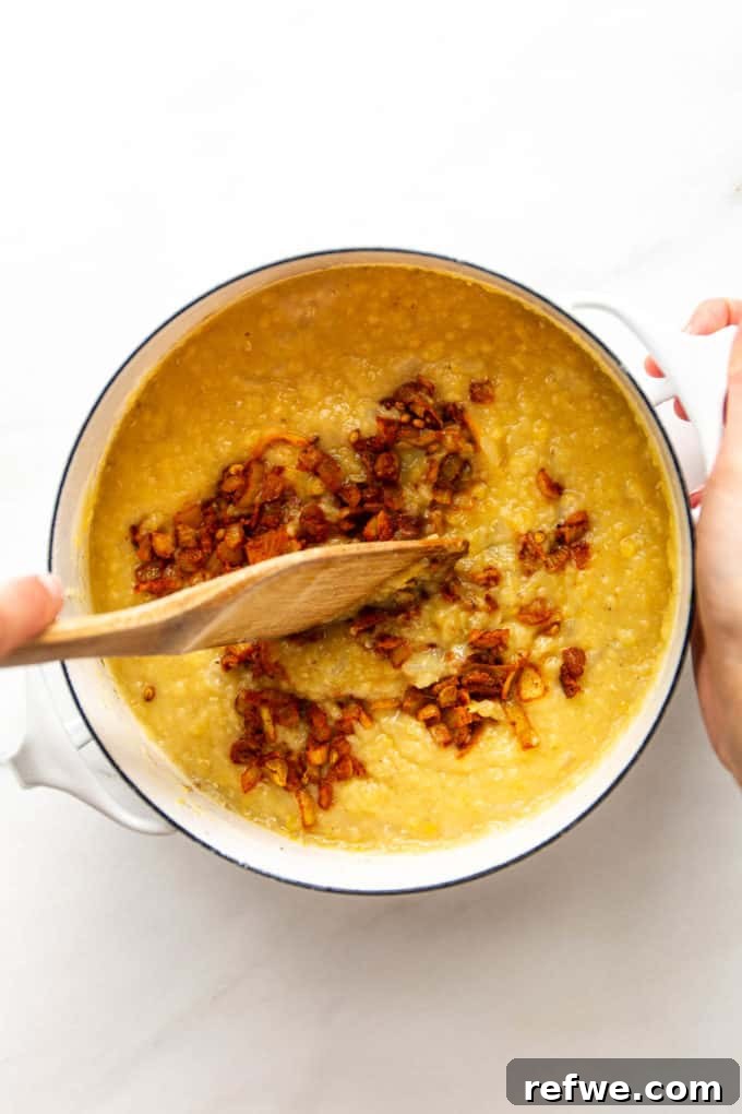 A wooden spoon stirring the spice mixture into the red lentil dahl in a white pot, combining all the flavors.