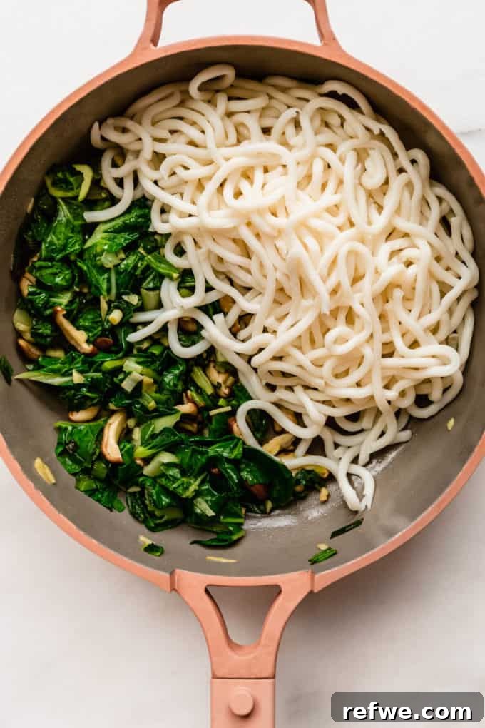 Fast and Simple Yaki Udon Stir Fry 6 Cooked udon noodles added to a pan with tender-crisp bok choy and shiitake mushrooms, ready to absorb the stir-fry sauce and create a flavorful dish.