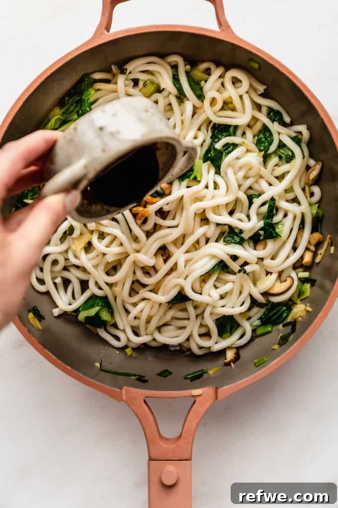 Fast and Simple Yaki Udon Stir Fry 7 Rich, dark stir-fry sauce being poured generously over udon noodles and vegetables in a pan, preparing for a thorough and even toss.