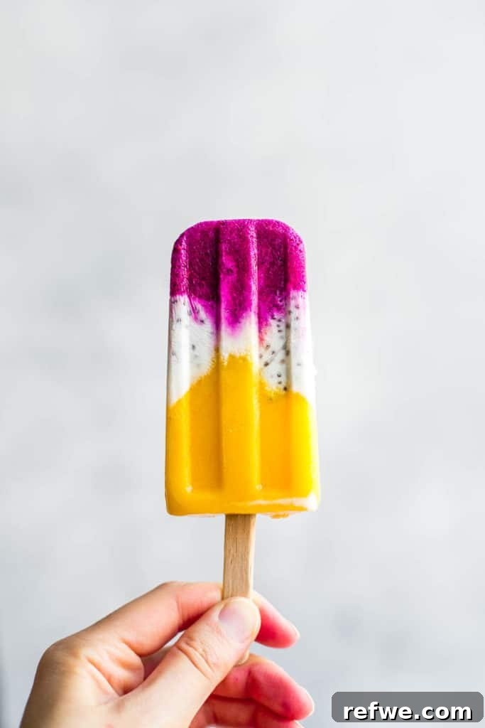 Exotic Coconut Chia Chillers 4 A close-up shot of a hand gently holding a vibrant Tropical Coconut Chia Popsicle, showcasing its beautiful layered colors of mango, coconut, and dragonfruit against a soft, blurred background.