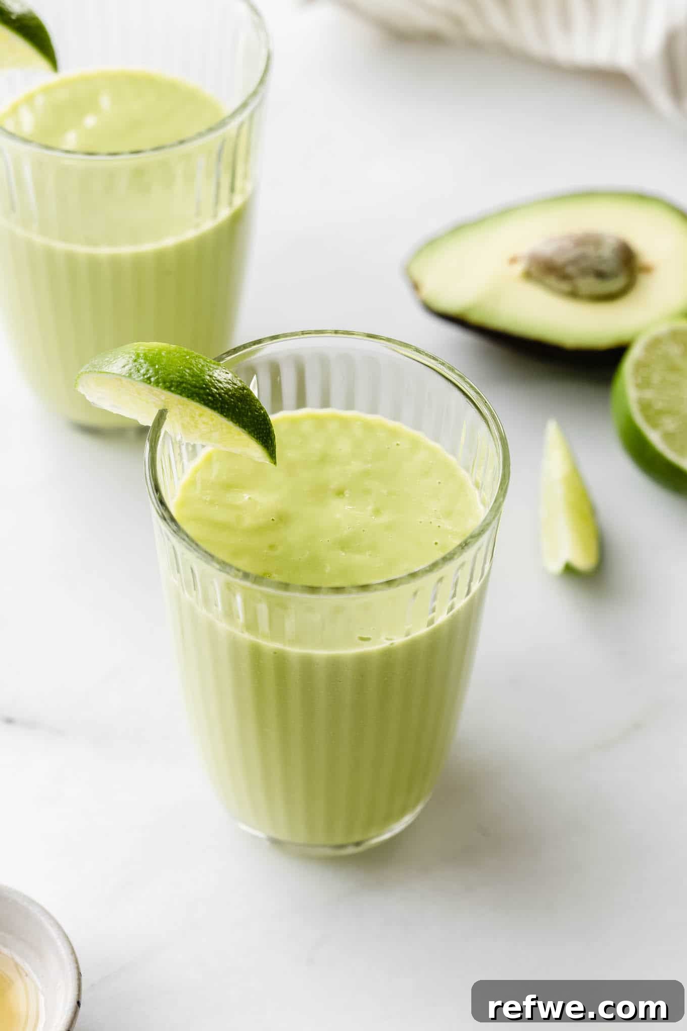 Tropical Green Dream 2 Two vibrant avocado mango smoothies on a marble counter, garnished with fresh avocado slices and lime wedges, ready to be enjoyed.