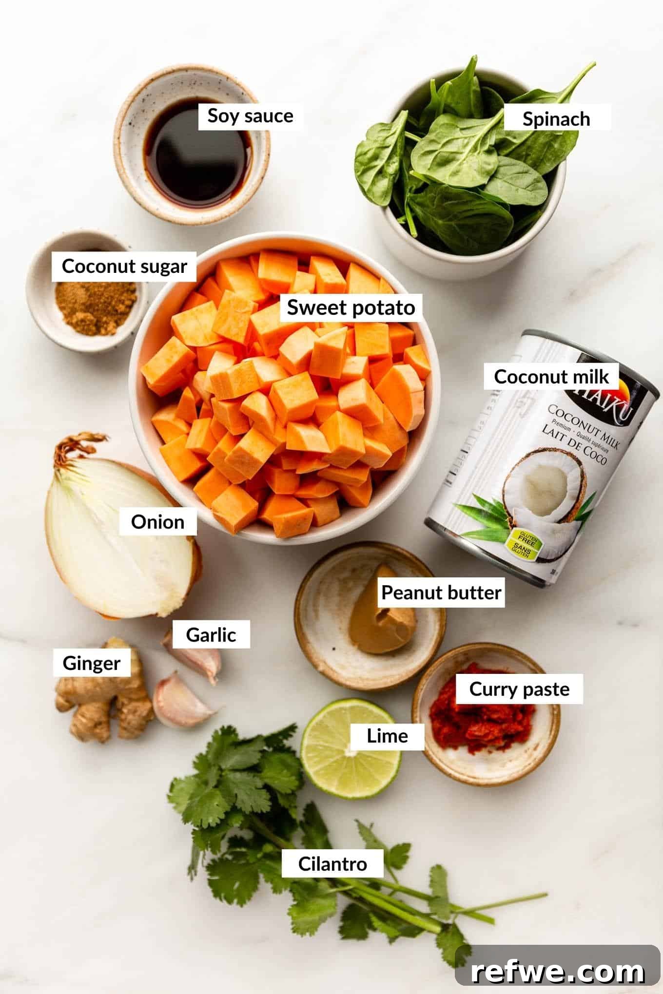A flat lay of fresh ingredients laid out on a white surface, including a sliced onion, ginger, garlic cloves, a jar of red curry paste, peanut butter, chili flakes, whole sweet potatoes, a can of coconut milk, vegetable broth, soy sauce, coconut sugar, lime, and spinach