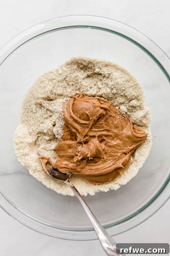 Funfetti Protein Bites 6 The process of combining dry almond flour and protein powder with creamy cashew butter in a clear mixing bowl, mid-stir.