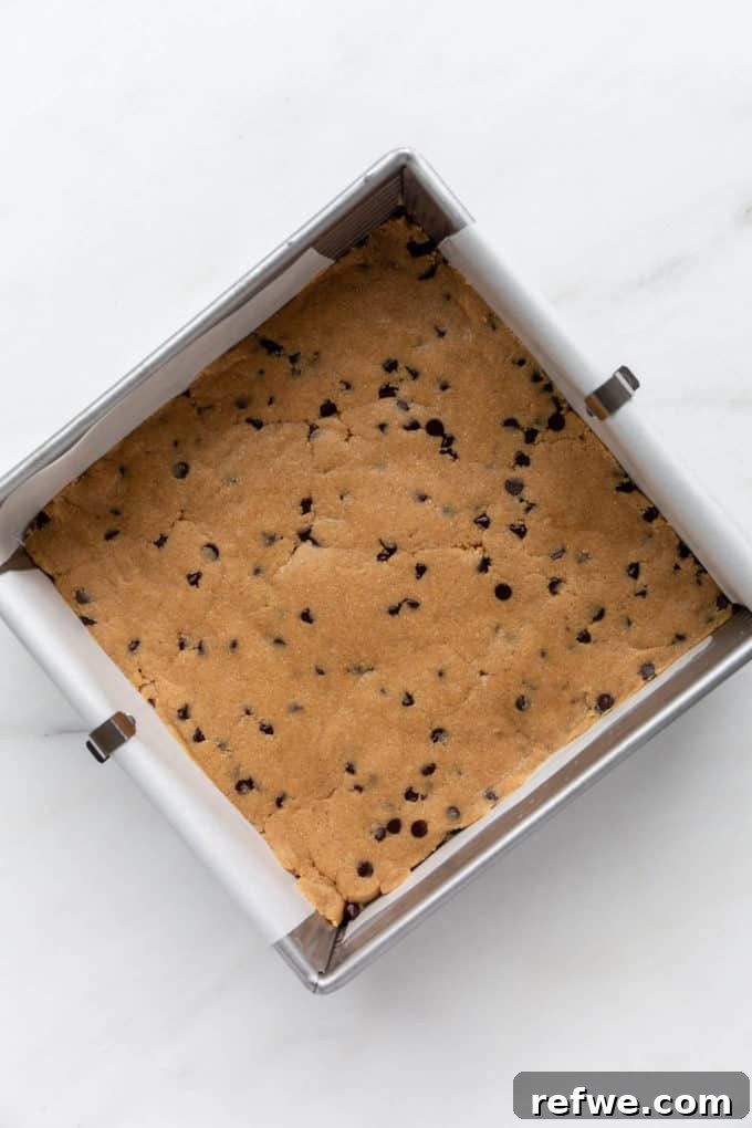 Fırınsız Sağlıklı Kurabiye Hamuru Barları 7 Smooth no-bake cookie dough is evenly pressed into an 8x8 inch pan lined with parchment paper, forming the base of the delicious bars.