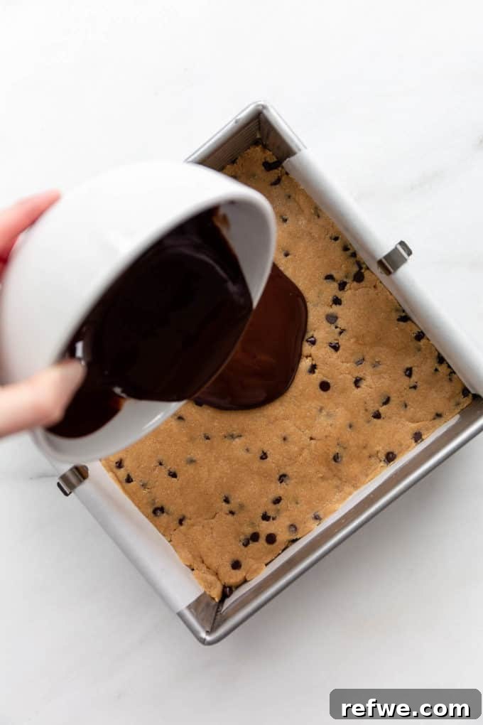 Fırınsız Sağlıklı Kurabiye Hamuru Barları 8 Rich melted dark chocolate, combined with coconut oil, is being poured over the smooth cookie dough layer in a baking pan, creating a luxurious topping.