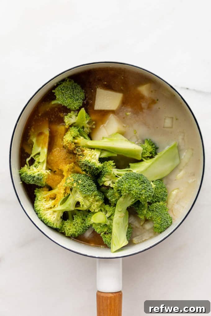 Velvety Vegan Broccoli Bliss 5 Broccoli florets, potatoes, coconut milk, and vegetable broth simmering in a white pot.