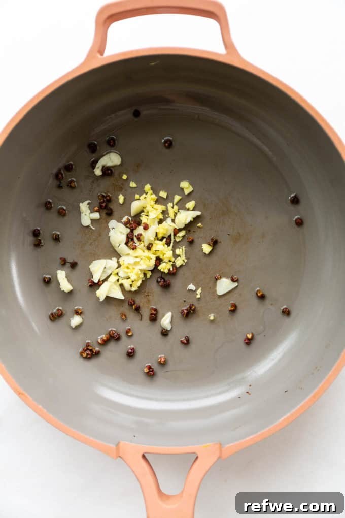 Fresh garlic, ginger, and Sichuan peppercorns being gently sautéed in a vibrant pink pan, infusing the oil with their potent and fragrant aromas.