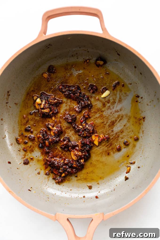 Rich, dark red fermented chili bean paste being stirred into the sautéed aromatics in a pink pan, creating the savory and spicy base of the Mapo Tofu sauce.