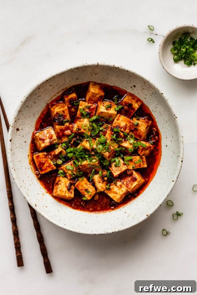 A vibrant bowl of Mapo Tofu, perfectly garnished with chopped green onions, with chopsticks resting nearby, signaling a delicious and authentic meal ready to be savored.